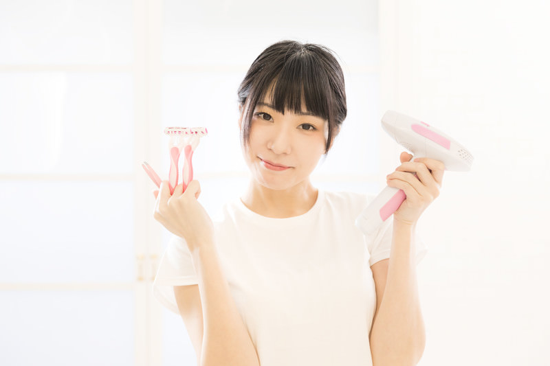 白いシャツを着たショートヘアの若い女性が右手にカミソリと左手に家庭用脱毛器を持っている